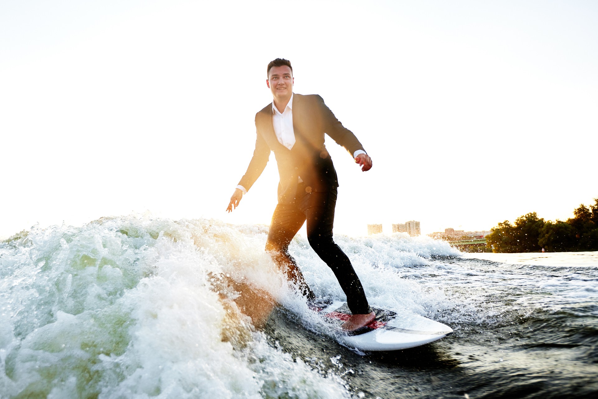 Young man in classic suit rides a wakeboard on the river or lake near city. Clerk escaped from a stuffy office to take up his favorite active sport. Best summer leisure after routine work. Sun flare.