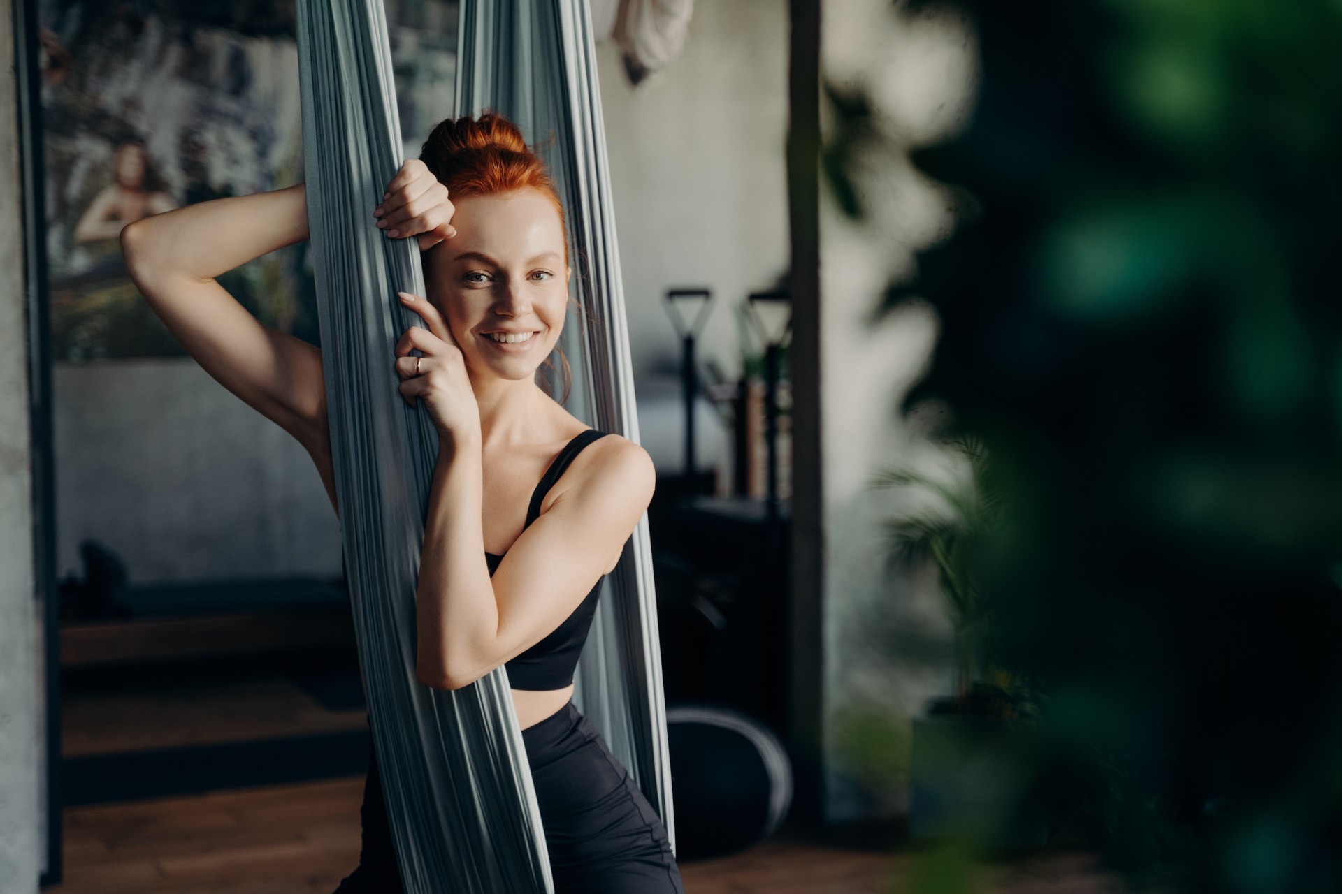 Young red haired woman sitting in hammock while visiting aerial yoga class